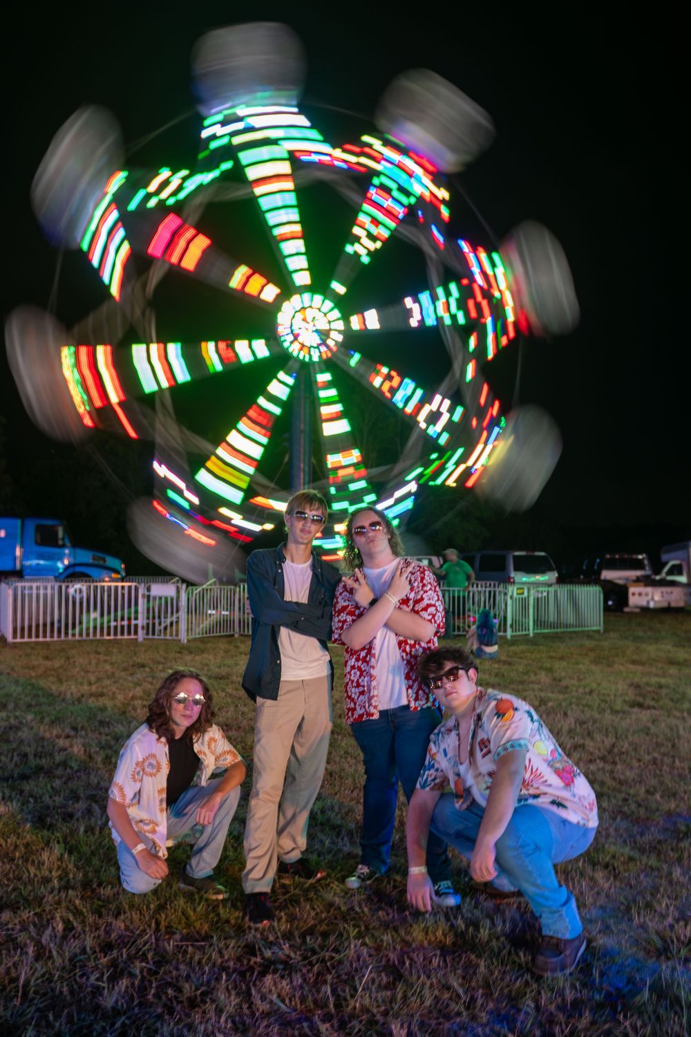 Long exposure photos show colorful carnival ride lights spinning in circles above people crouching in a grassy field at night.