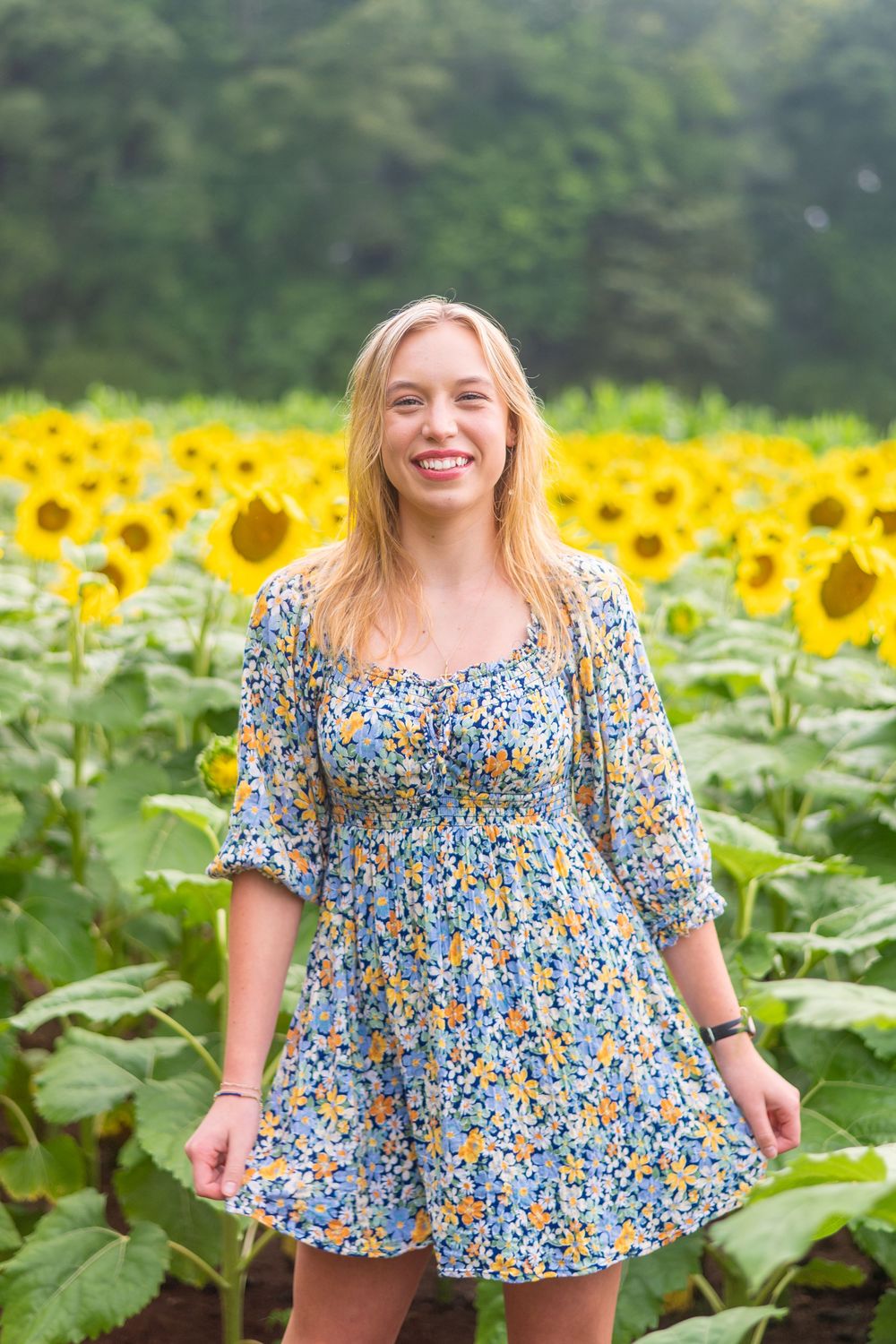 A person in a blue floral dress poses joyfully in a vibrant field of blooming sunflowers on a summer day.