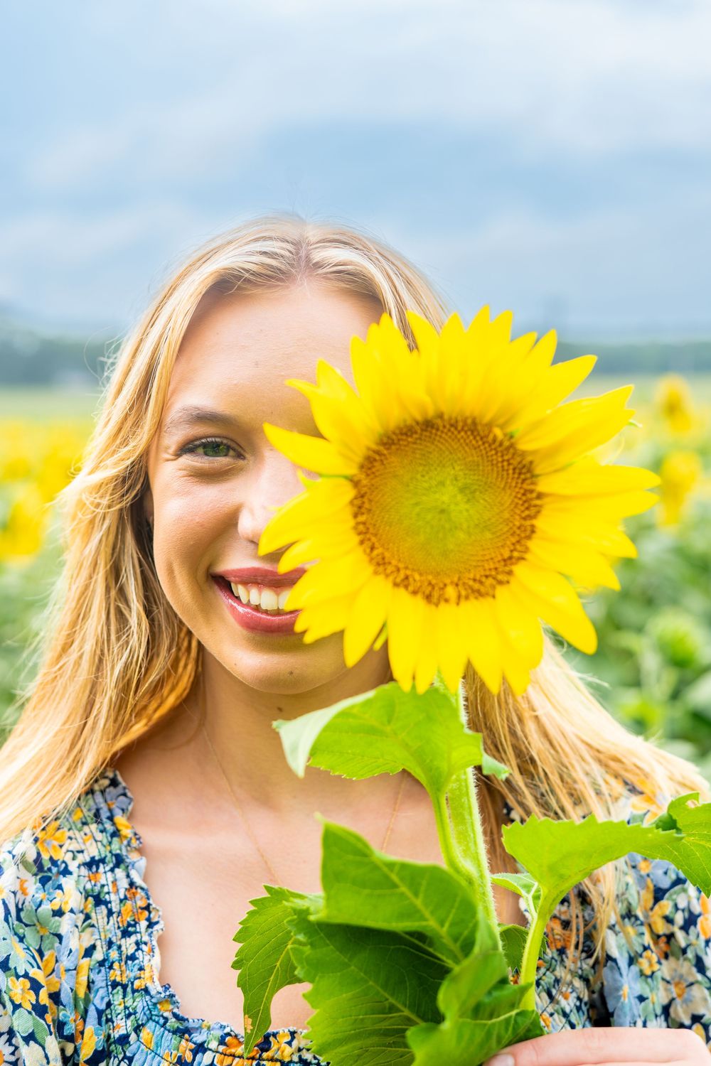 A bright yellow sunflower stands tall in a sunny field while someone in a blue floral dress poses behind it.