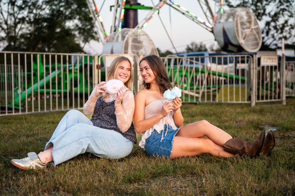Two friends laugh together while relaxing on grassy hill during summer photoshoot.