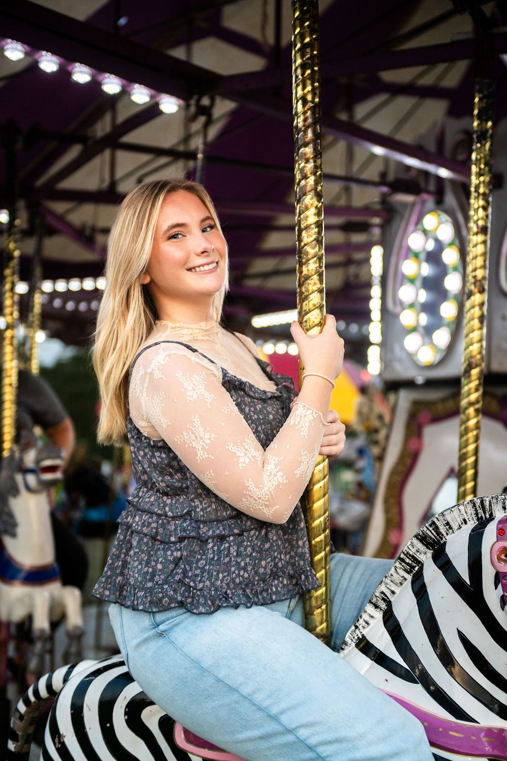 Person riding on a carousel with zebra-striped horse at an amusement park with colorful lights in the background.