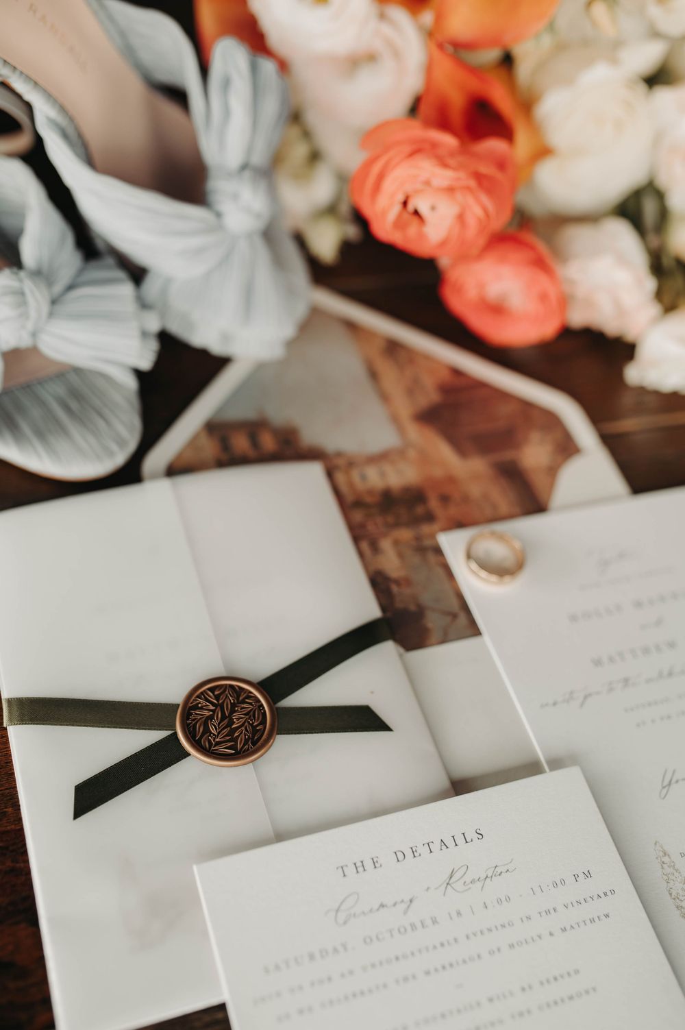 Wedding invitation suite featuring a bronze wax seal, olive green ribbon, and coral floral arrangement nearby.