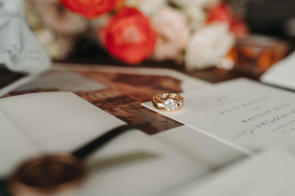 Close-up of gold wedding rings resting on a wedding invitation with a floral bouquet in the background.