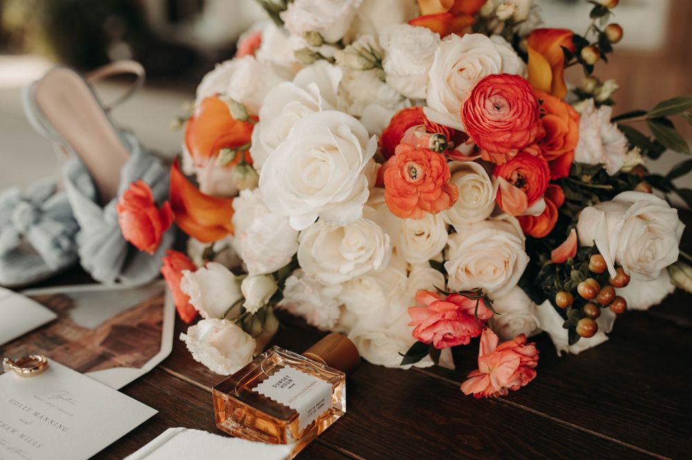 Vibrant bridal bouquet of white roses and coral ranunculus beside a perfume bottle on a wooden table.