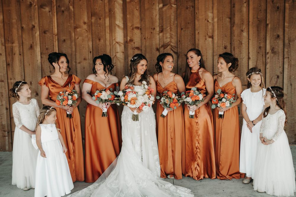 Bridal party in burnt orange satin gowns with flower girls posing in front of a rustic barn wall.