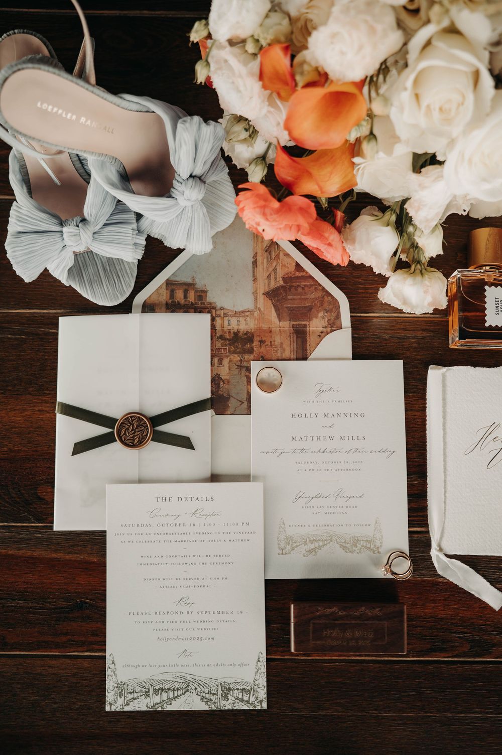 Elegant wedding flat lay with floral invitations, wax seal, blue heels, and orange and white bouquet on wood.