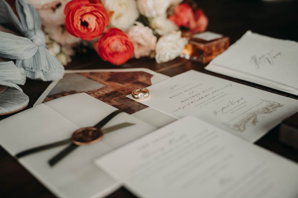 Close-up of wedding invitation suite with wax seal, rings, and coral ranunculus bouquet on dark wood table.