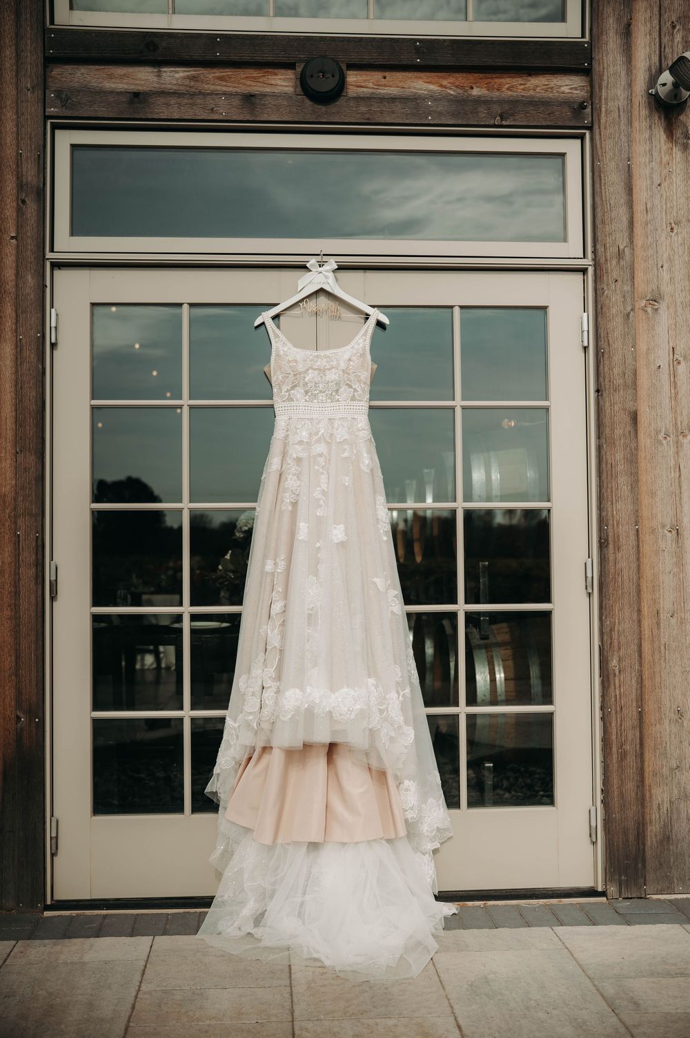 Floral lace wedding gown with layered skirt hanging on glass barn doors in natural light.
