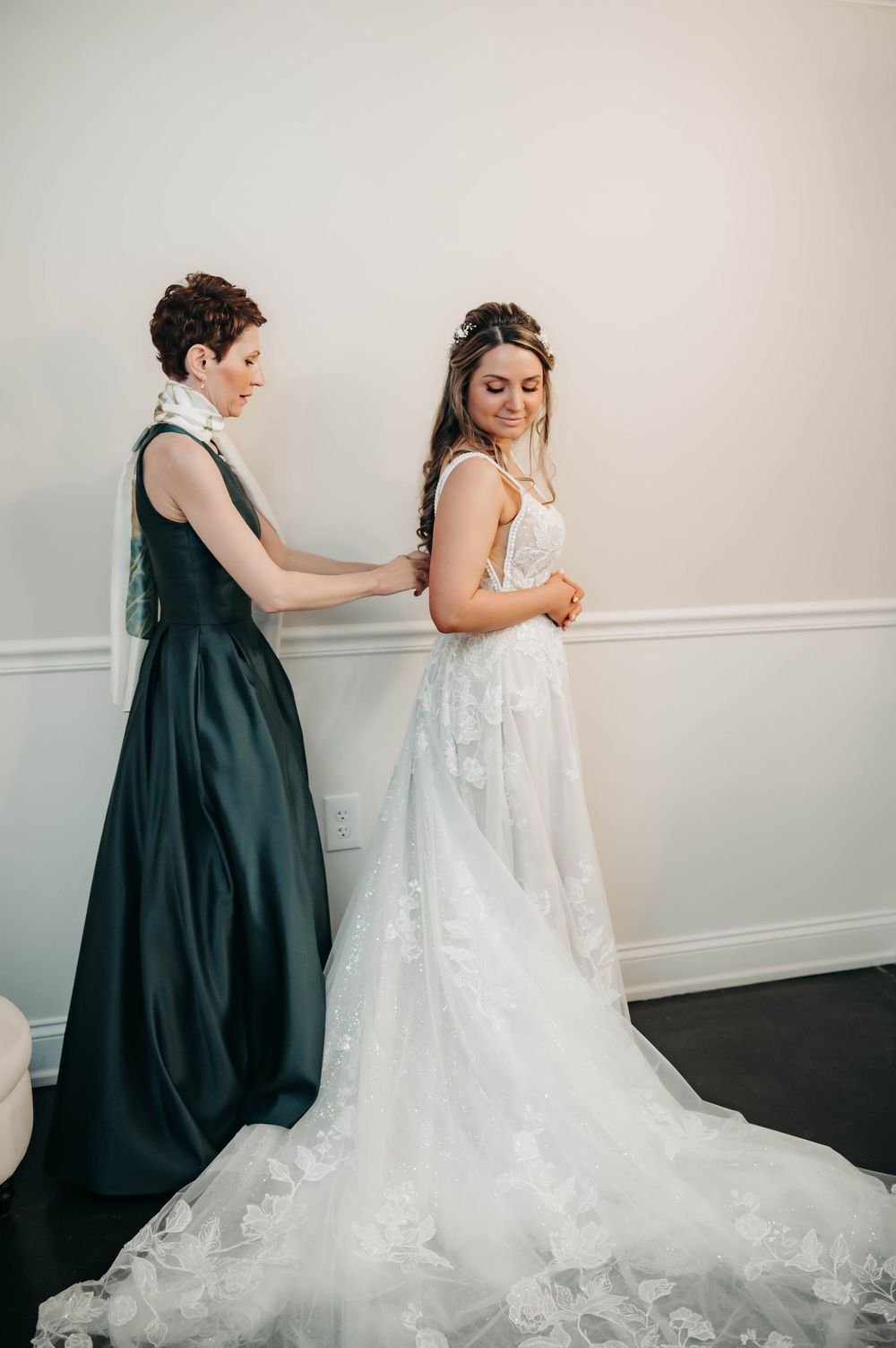 Bridesmaid in dark green gown helping bride fasten her lace wedding dress with long train before ceremony.