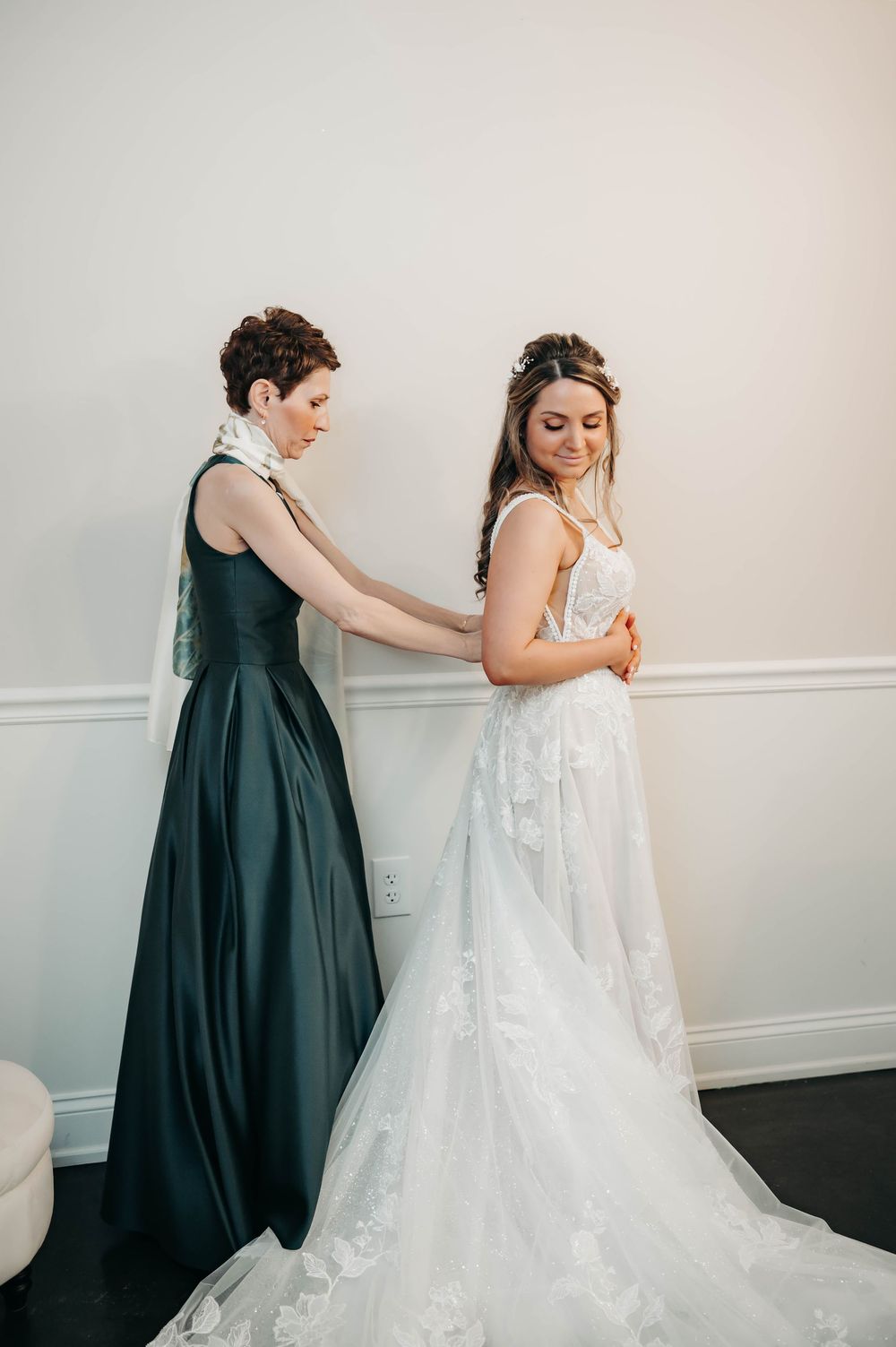 Woman in elegant dark green dress carefully buttoning the back of a brides lace wedding gown with cathedral train.