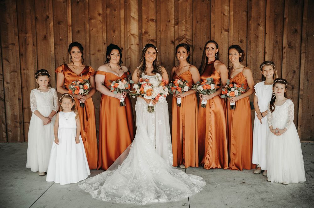 Bride with bridesmaids in burnt orange satin gowns and flower girls in white lace at rustic barn.