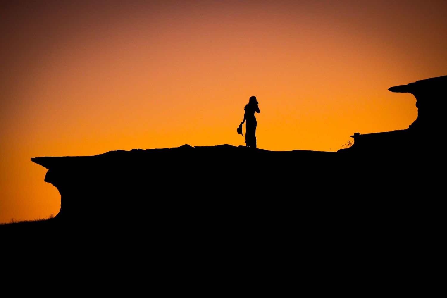 A silhouetted figure stands on a rocky outcrop against a vibrant orange sunset sky.