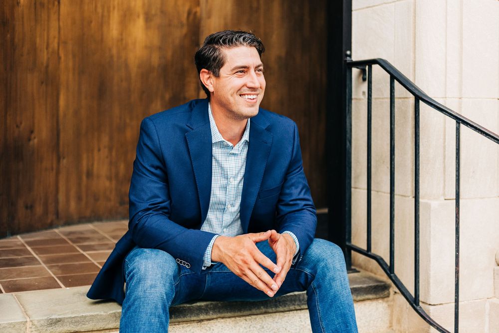 A person in a navy blazer and jeans sits casually on outdoor steps with a warm smile against a wooden door background.