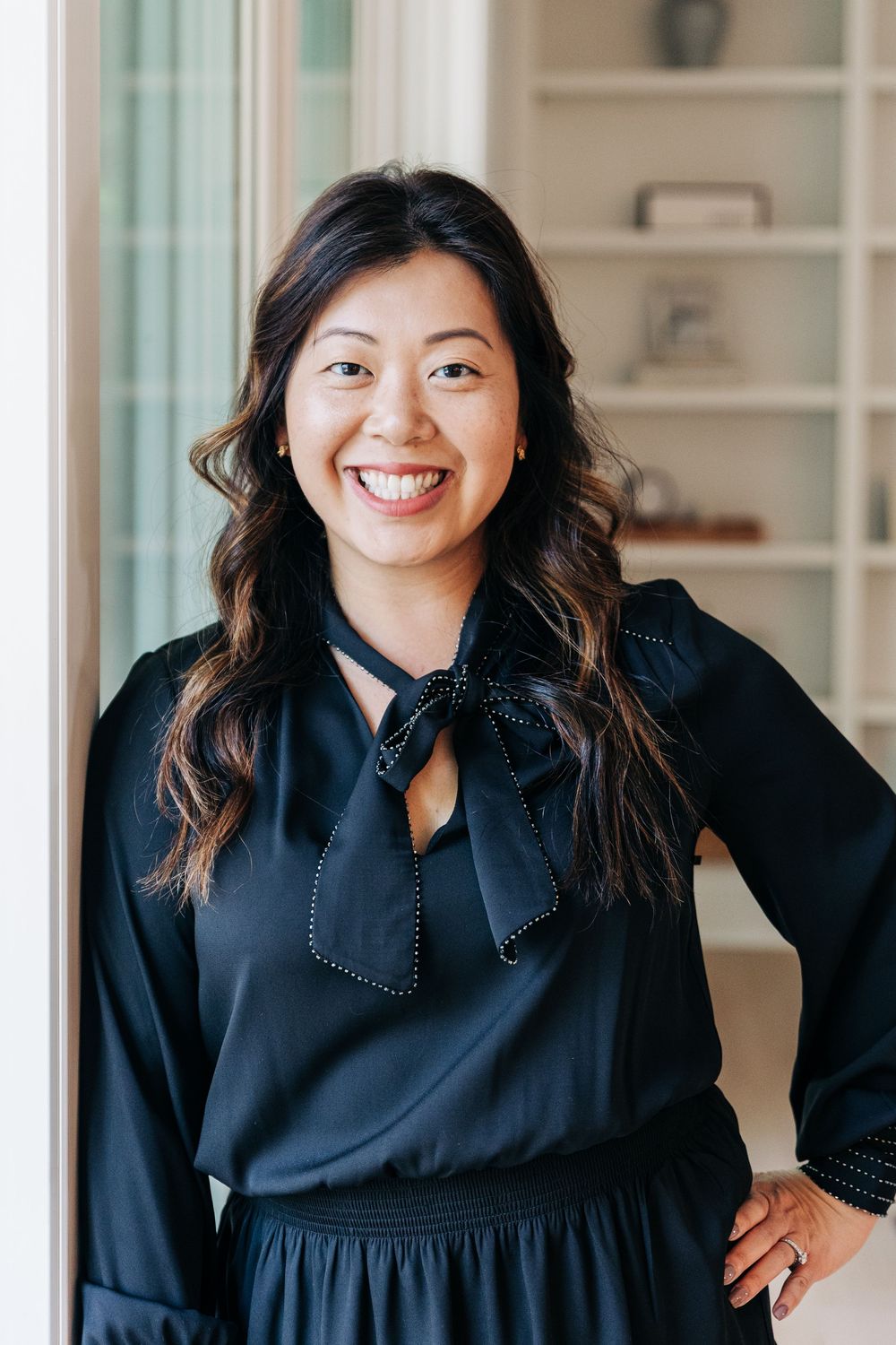 A person wearing a black dress with polka dot details poses and smiles in a bright indoor setting.