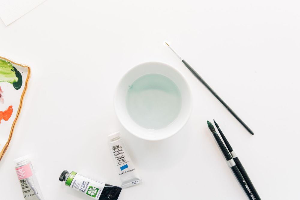 Flat lay photography setup with white desk, paint brushes, watercolor supplies and art materials.