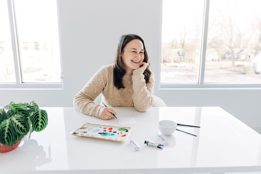 Woman in beige sweater smiles while working at bright modern desk with laptop and office supplies.