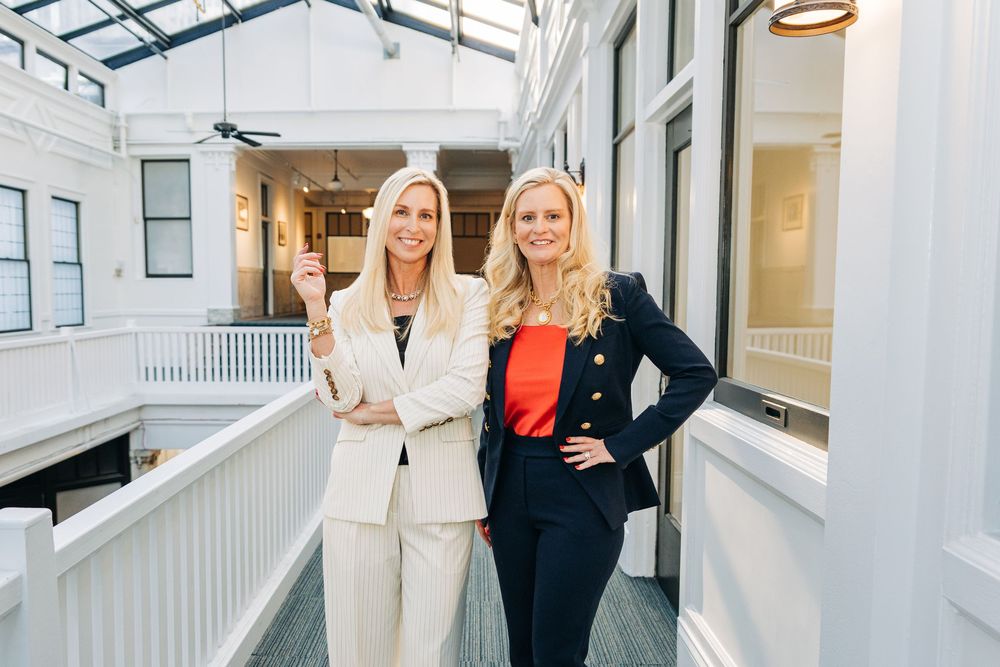 Two people in professional business attire stand together in a bright modern office hallway.