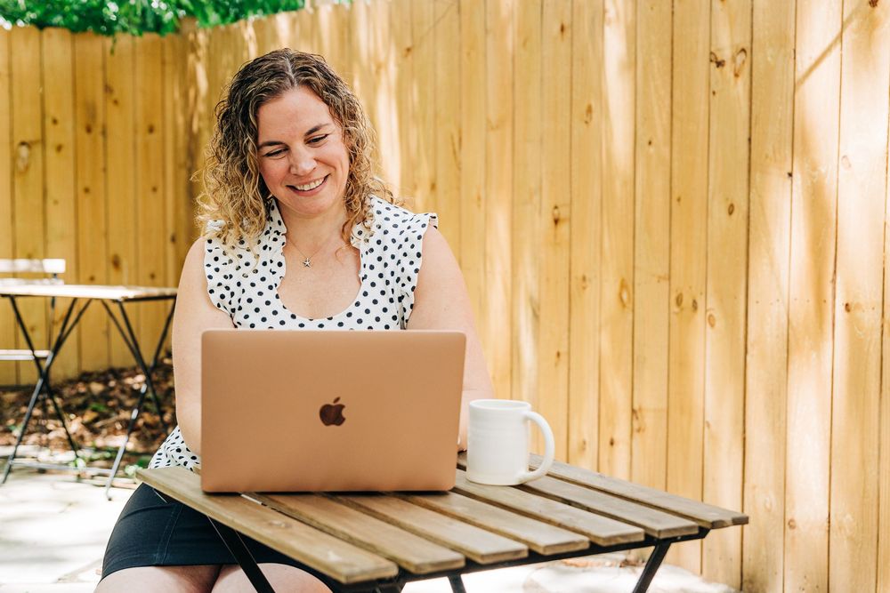 Woman working on laptop outdoors on wooden deck with coffee mug and greenery in background.