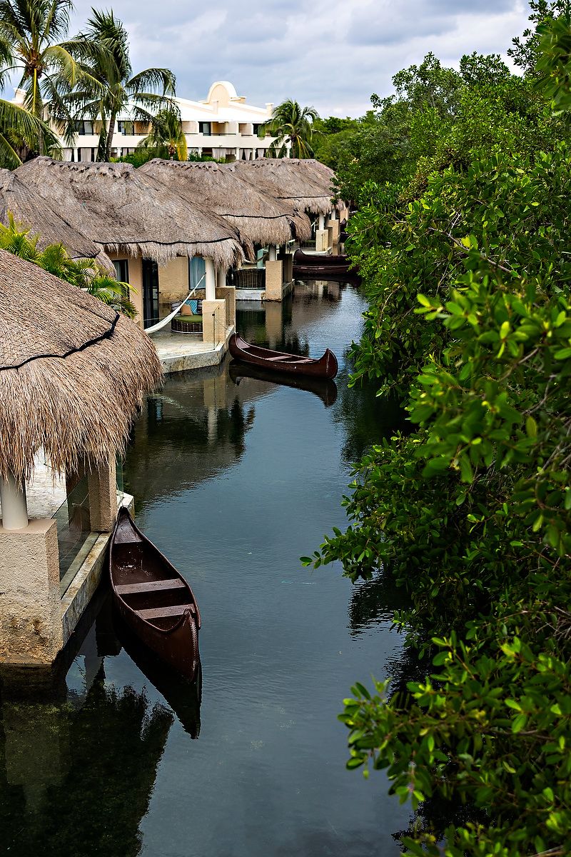 Thatched-roof waterfront bungalows with wooden canoes line a tranquil canal at a tropical resort surrounded by palm trees.