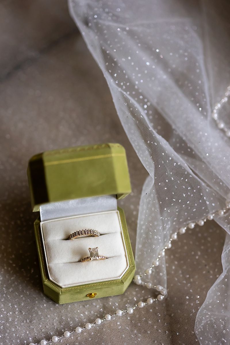 Two delicate engagement rings rest in a sage green velvet ring box next to sheer dotted tulle fabric.