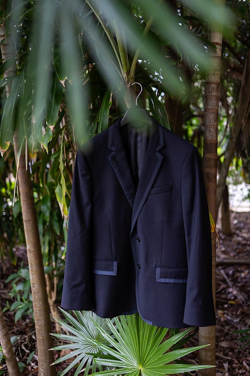 Three black formal suit jackets hanging on hangers among tropical palm fronds and bamboo in an outdoor setting.