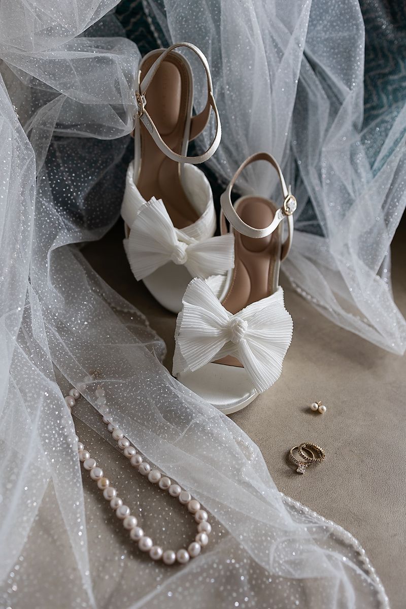Wedding accessories including white bridal shoes, delicate tulle veil, and pearl necklace arranged on light surface.