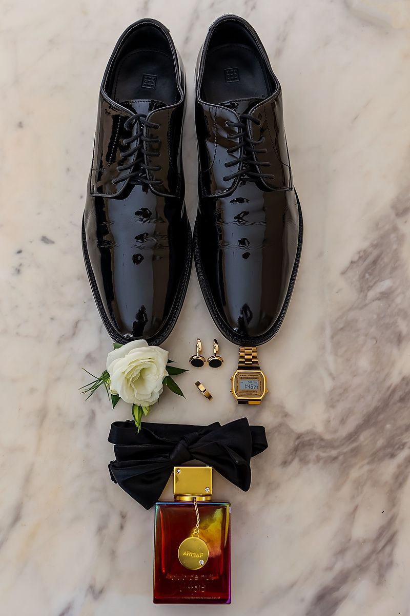 Black formal dress shoes with bow tie, white rose boutonniere, and gold accessories arranged on marble surface.