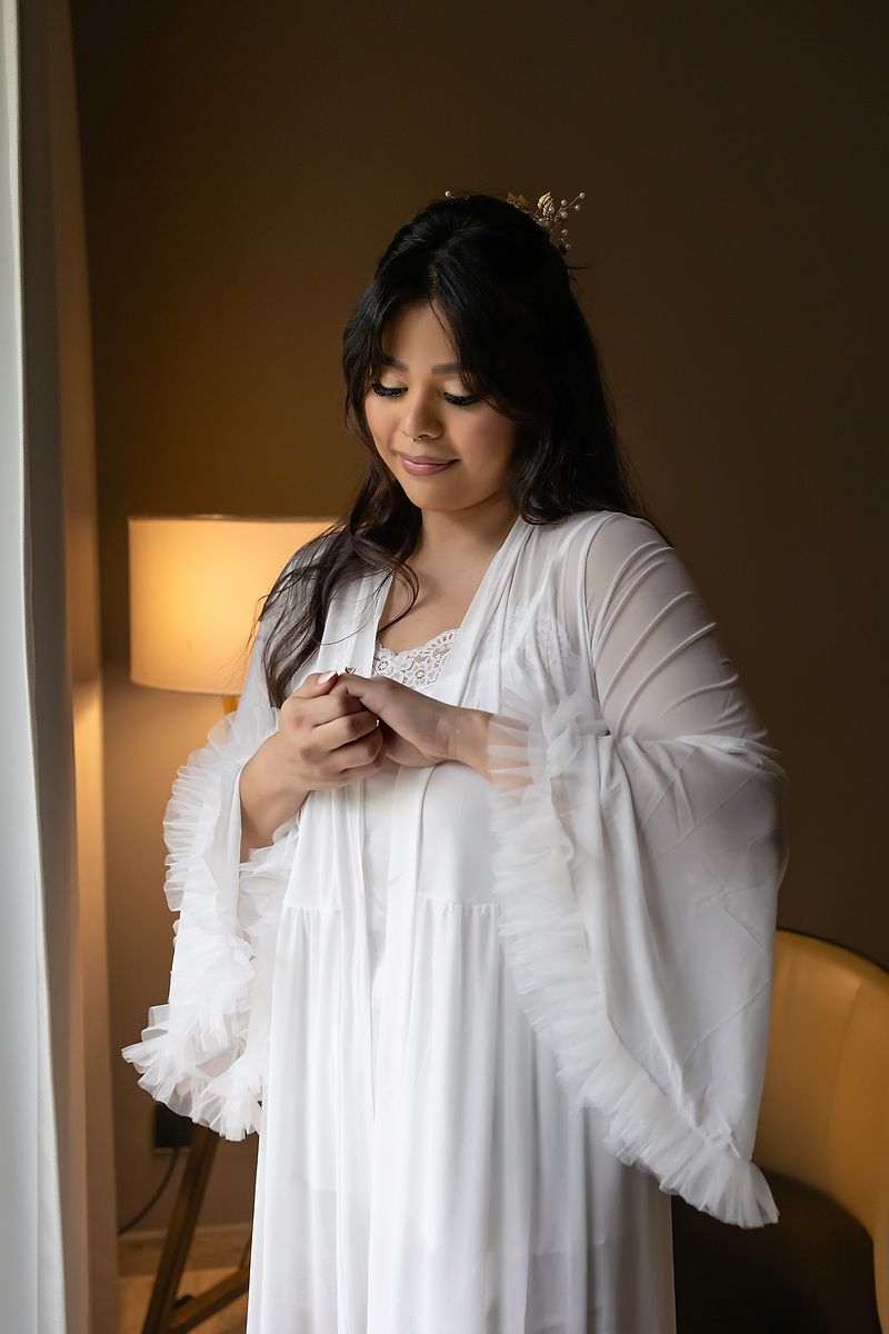 A person in a white ruffled robe stands near a window with warm lighting from a bedside lamp.