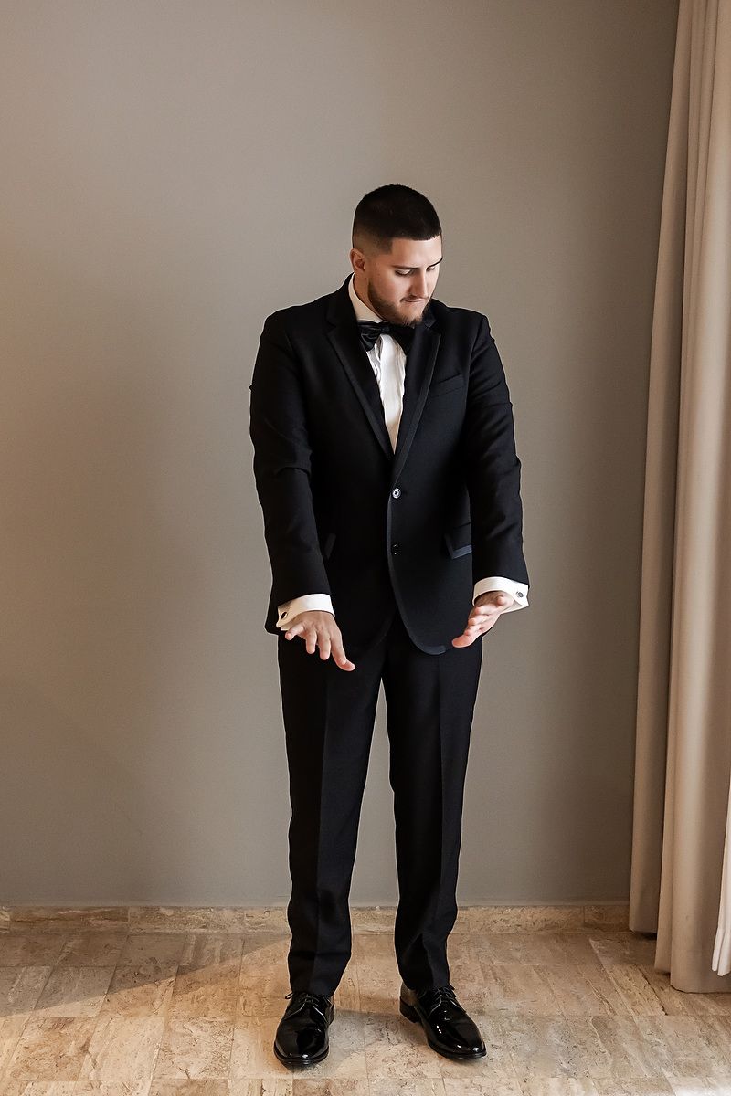 A person wearing a classic black tuxedo and bowtie adjusts their sleeve while standing in a room.