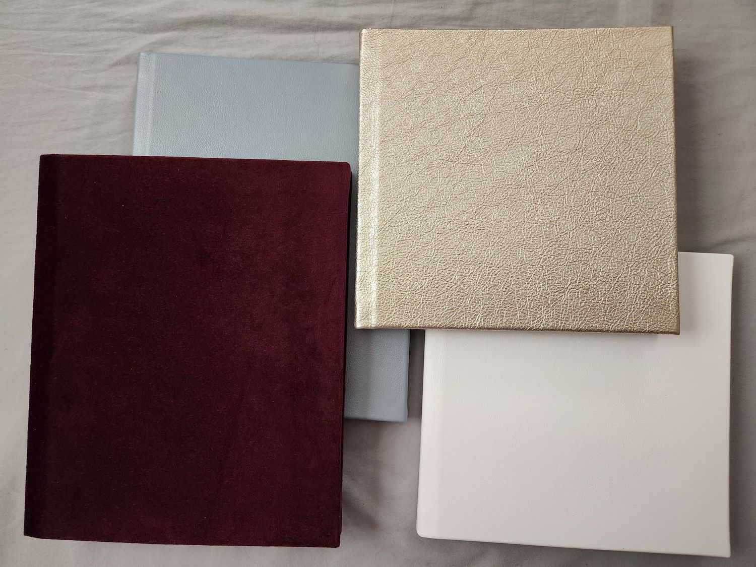 Boudior Albums. Four square fabric or material samples in burgundy, cream, light gray and white colors arranged on a surface.