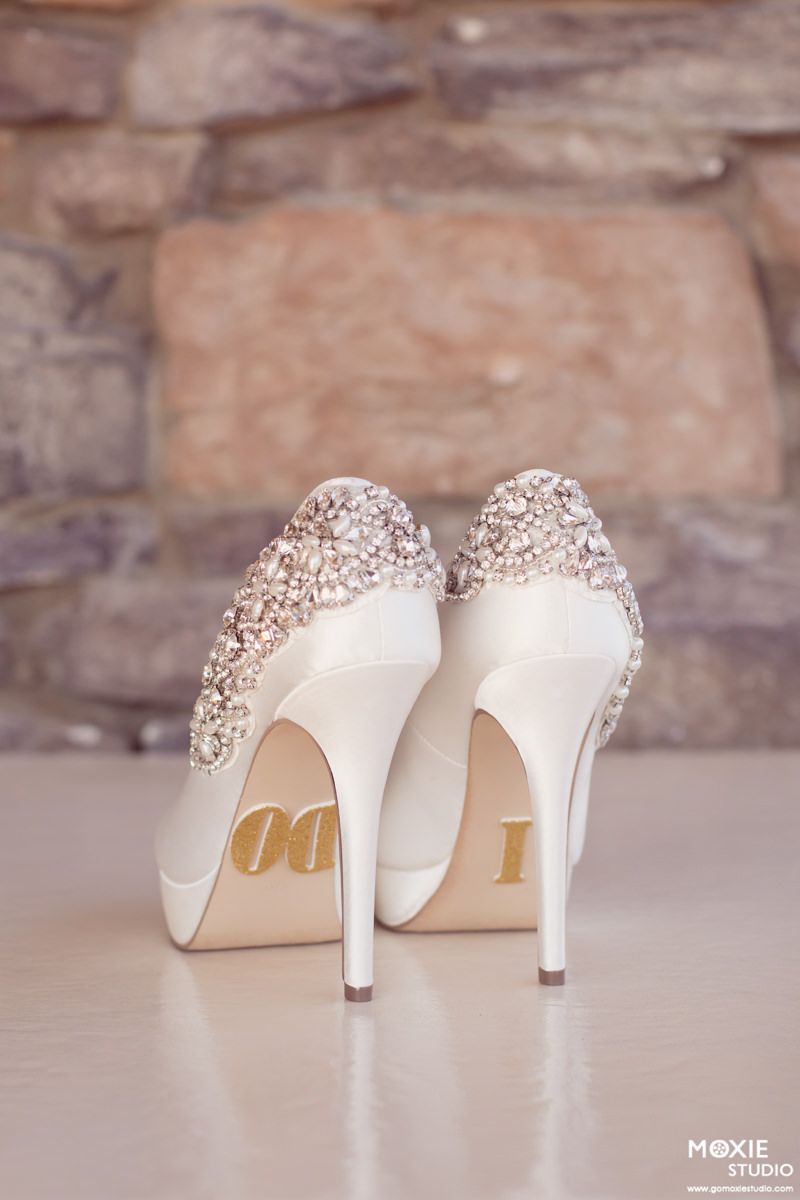 Elegant white bridal shoes with crystal embellishments on the heel shown from different angles against a stone wall backdrop.