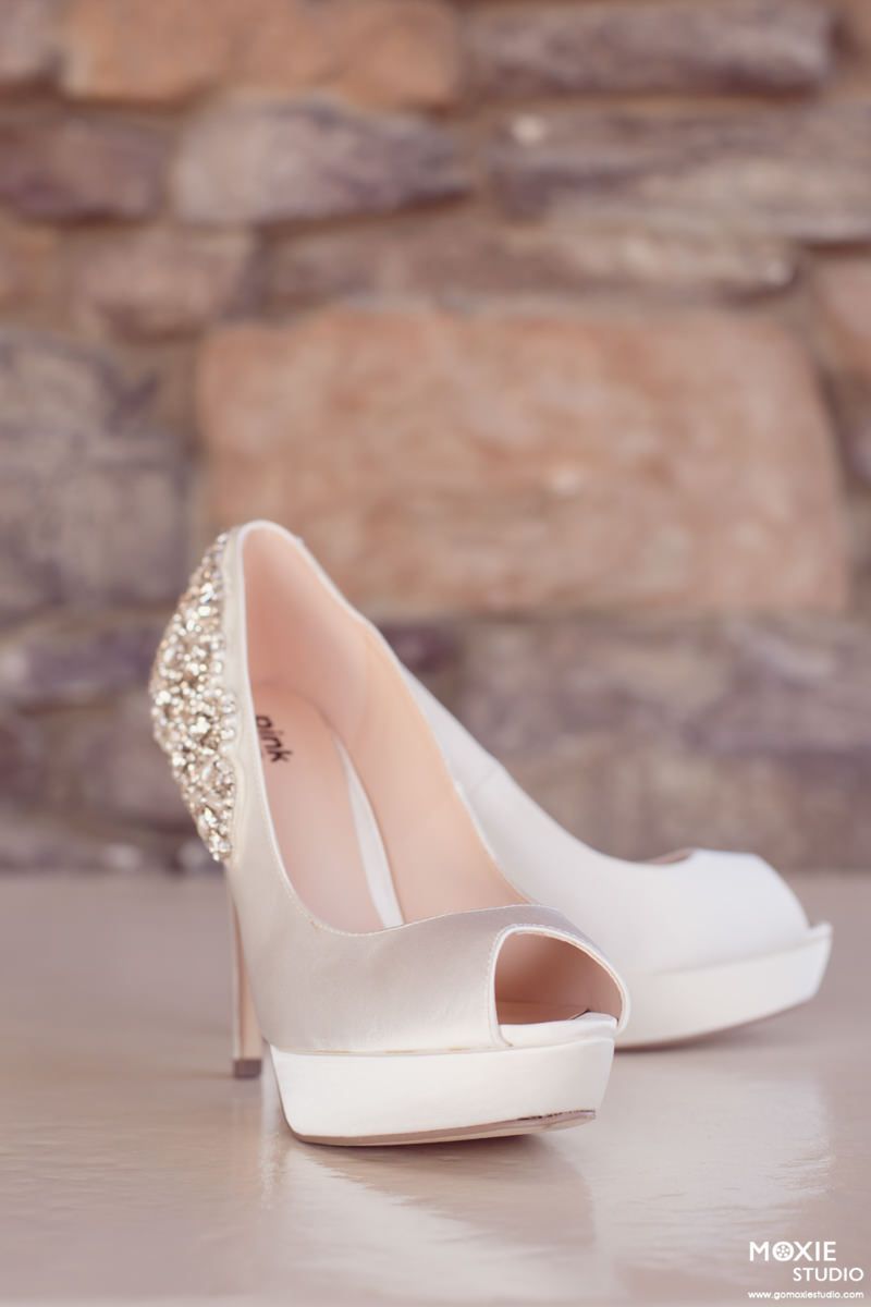 White satin bridal shoes with crystal embellished heels shown in sequence against a rustic stone wall background.