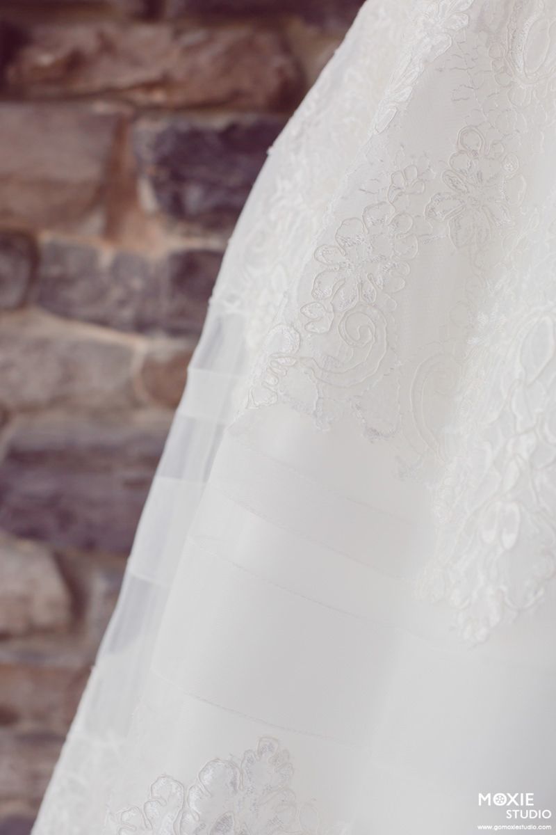 Close-up detail of delicate lace pattern on white wedding dress fabric against rustic stone wall background.
