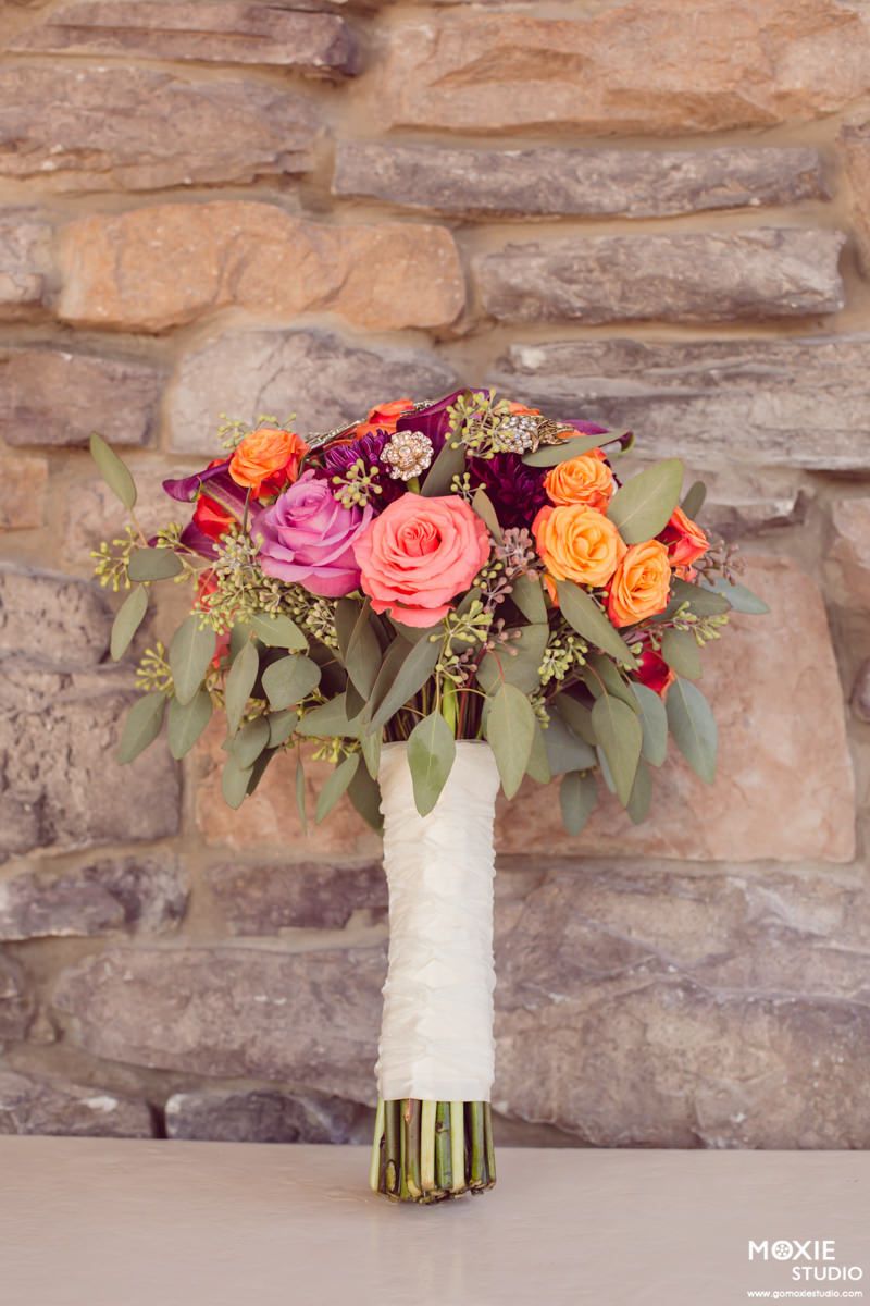 Vibrant bridal bouquet with pink, orange and burgundy roses wrapped in white lace ribbon against stone wall backdrop.