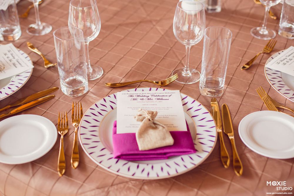 Formal place setting with gold cutlery, wine glasses, and pink napkin on rose gold tablecloth.