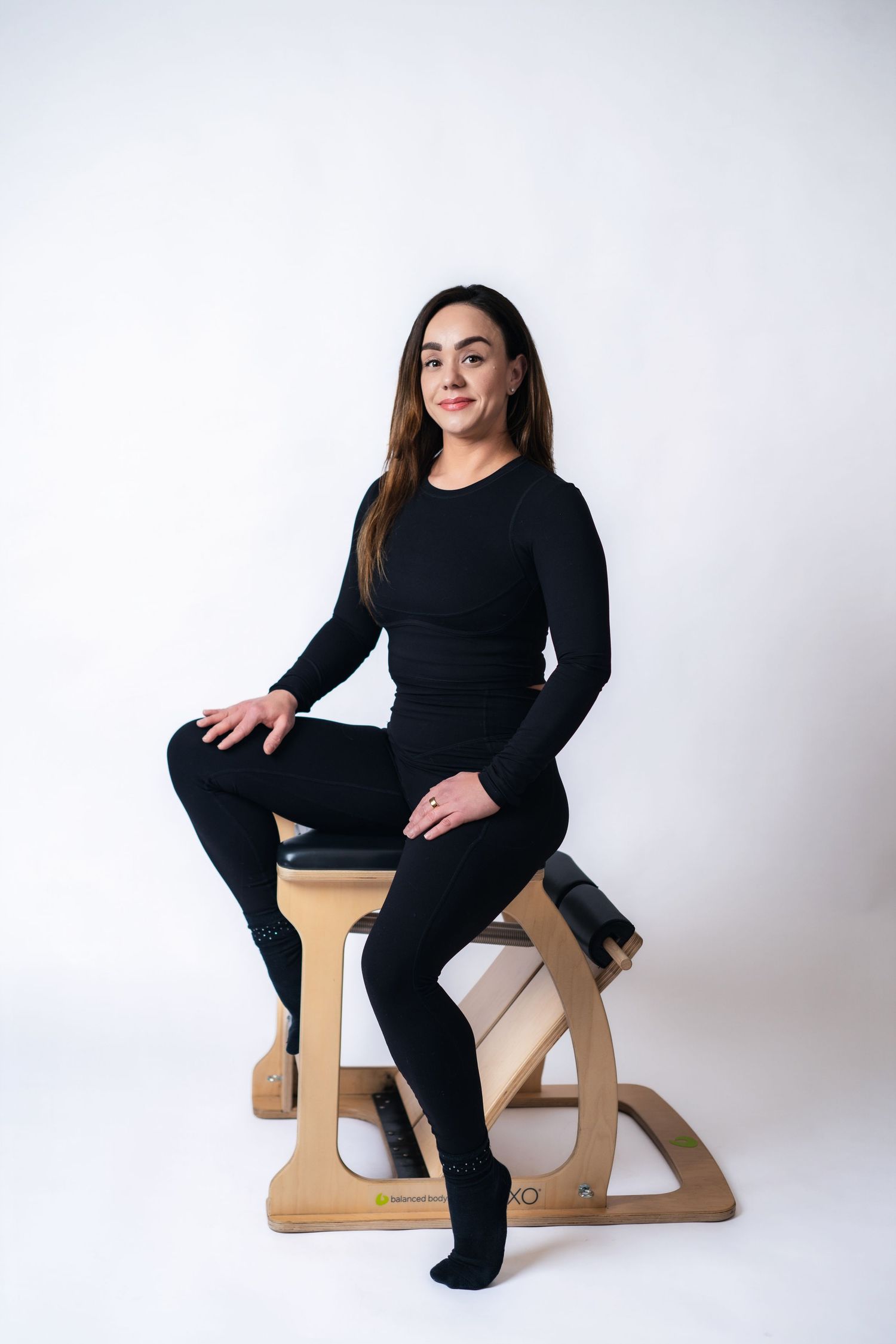 Person wearing all black athletic wear sits on a modern wooden ergonomic kneeling chair against white background.