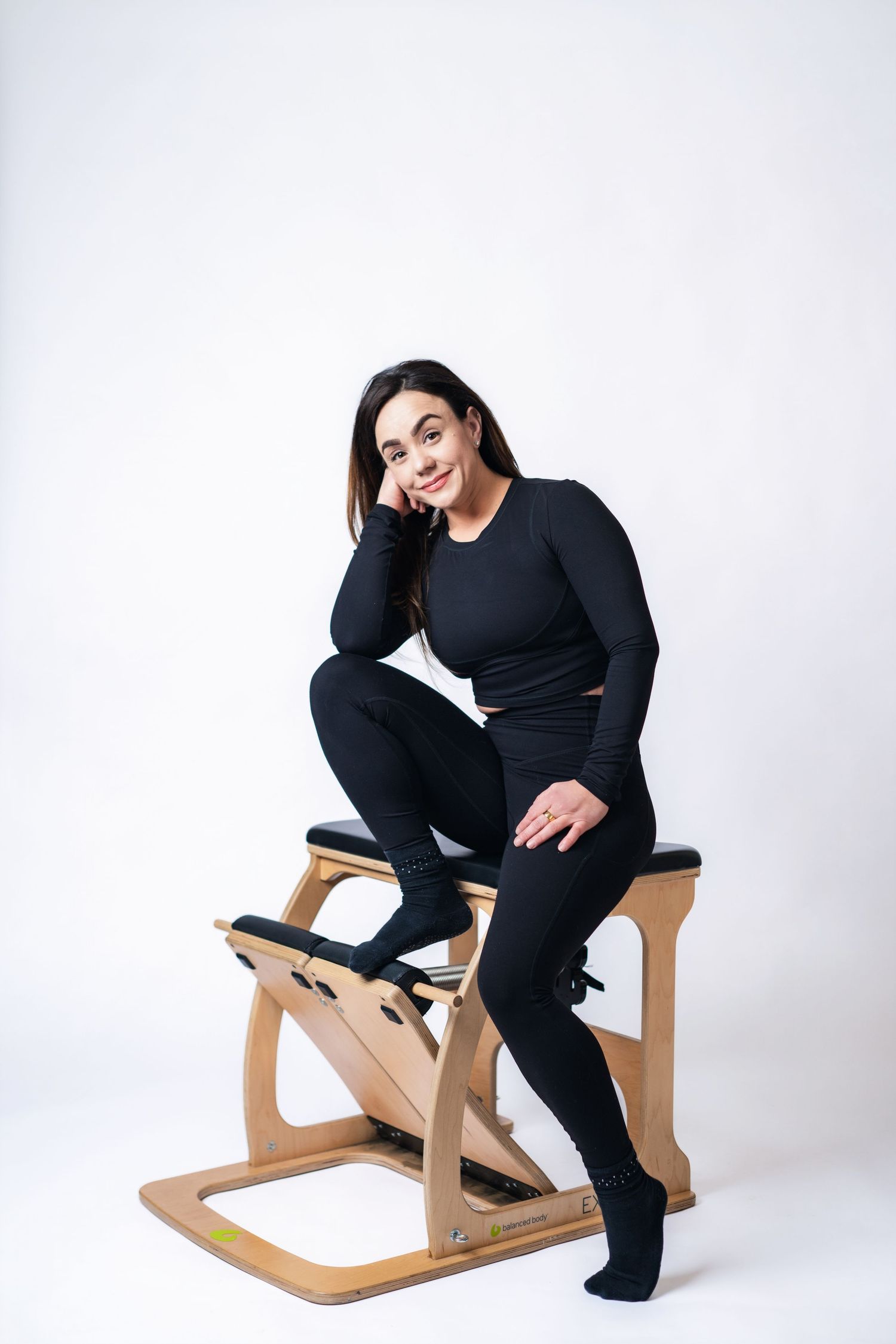 Person wearing black outfit sits in various poses on a modern wooden kneeling chair against white background.