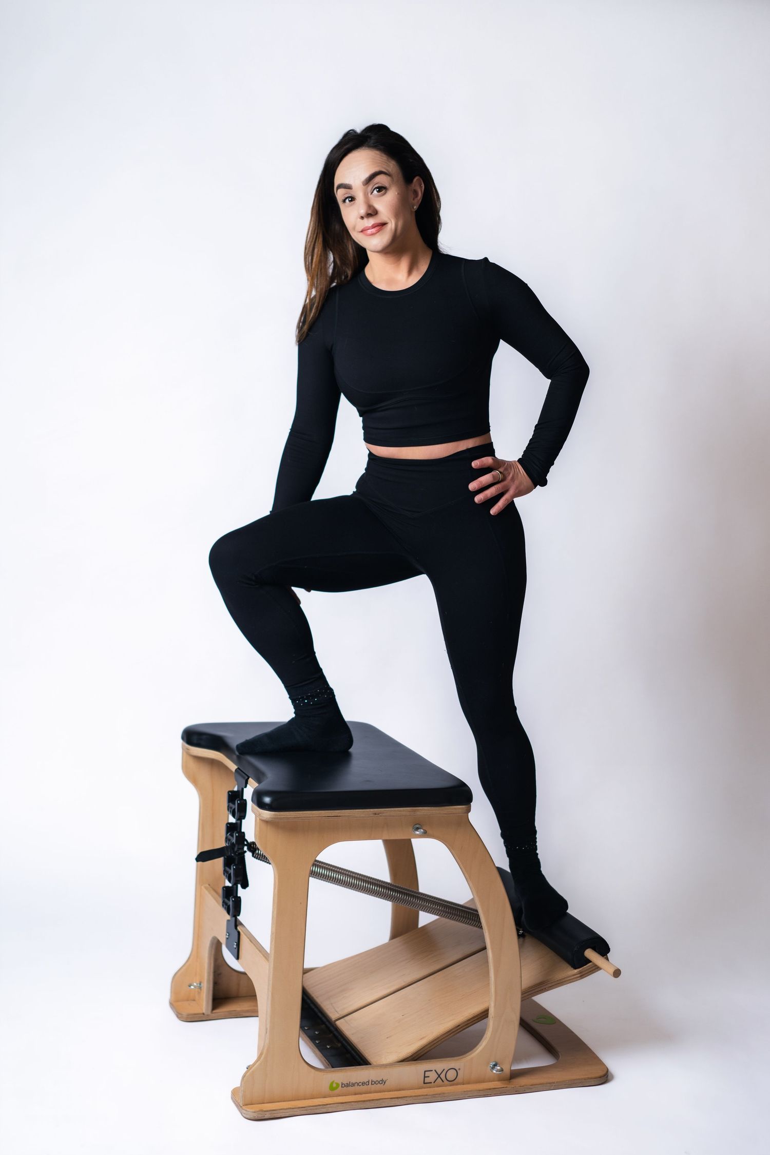 Athletic figure in all-black workout attire poses on wooden exercise equipment against white background.