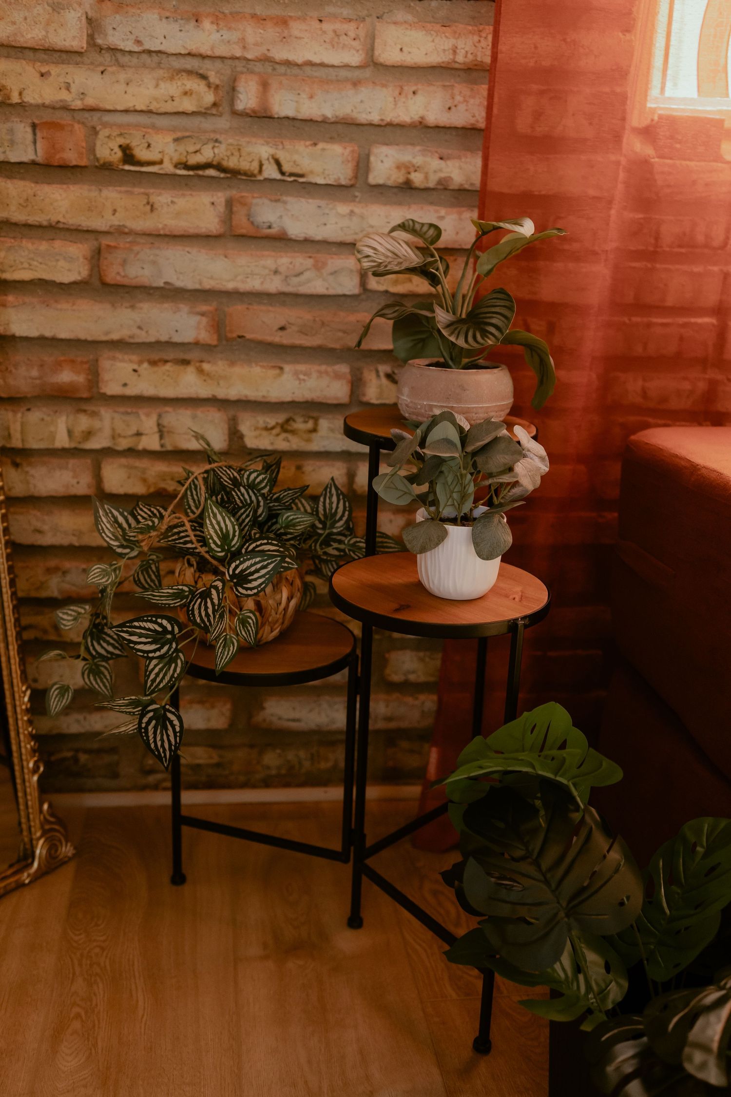 Small round side tables with black metal legs and wooden tops sit against a brick wall with potted plants in warm lighting.