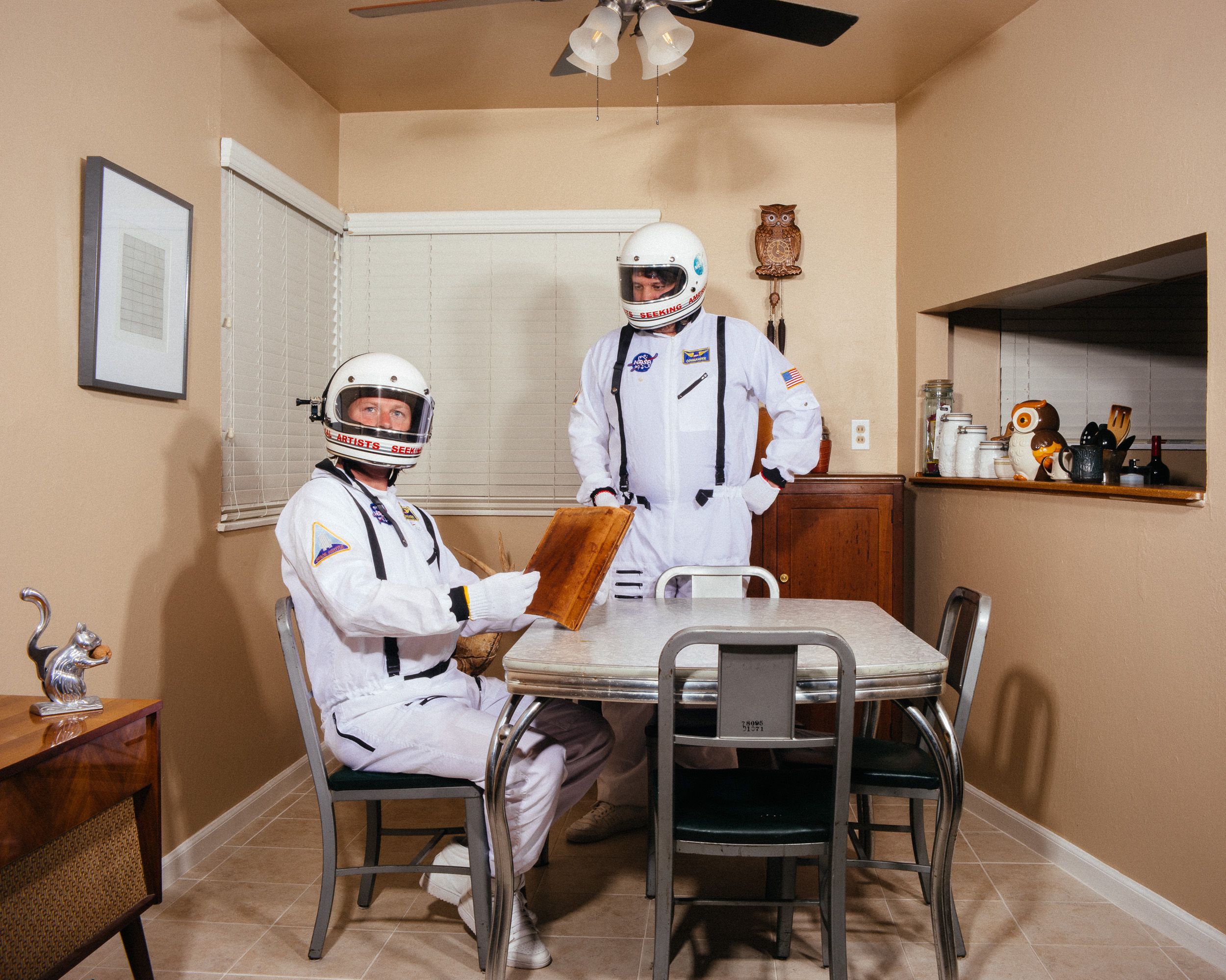 Two astronauts in white spacesuits sit at a small dining table in a beige-colored room.
