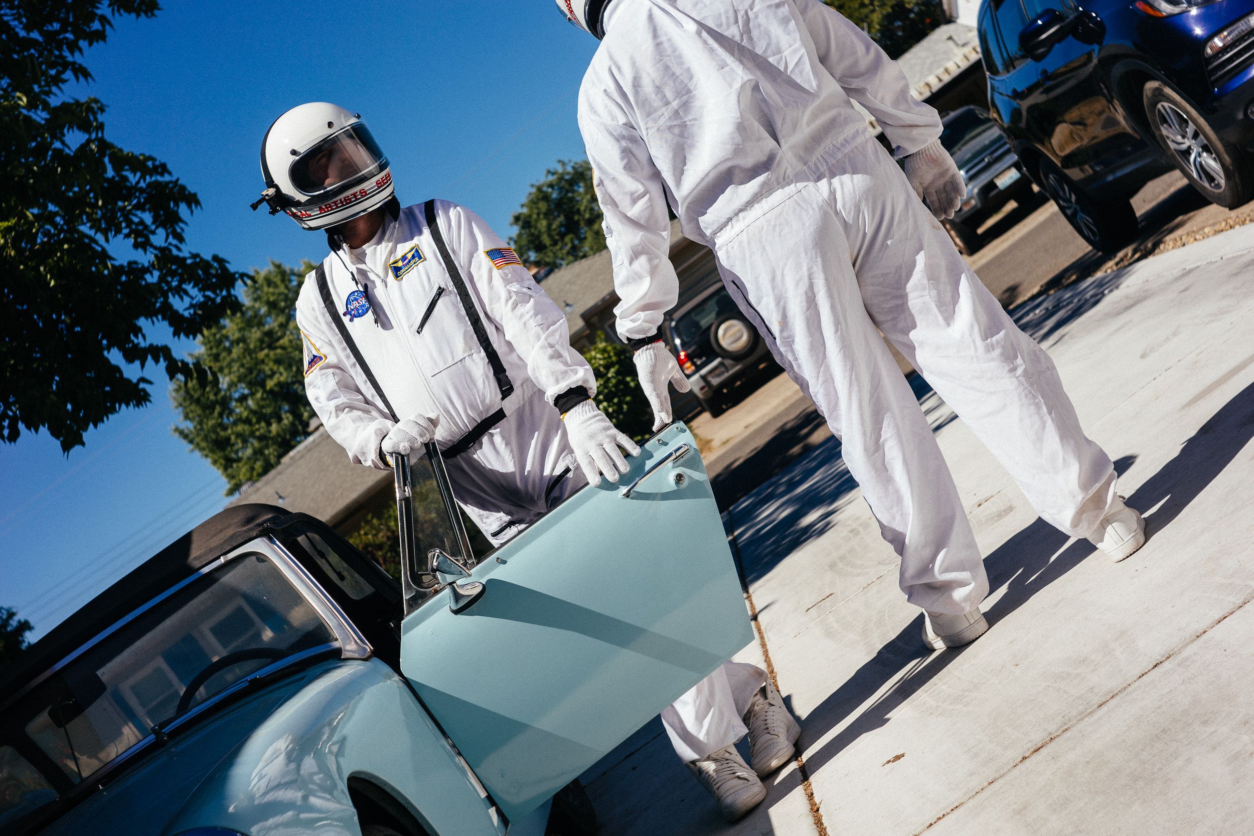 A person in white protective gear inspects a light blue vehicle in an outdoor setting during a sunny day.