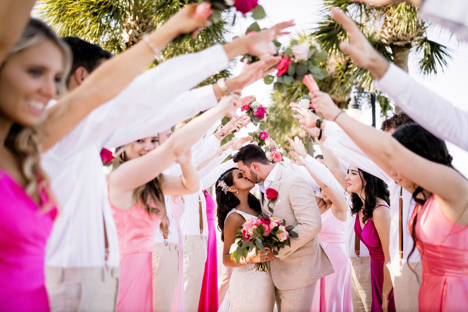 Wedding party raises floral bouquets to form an arch as bride and groom share a kiss.