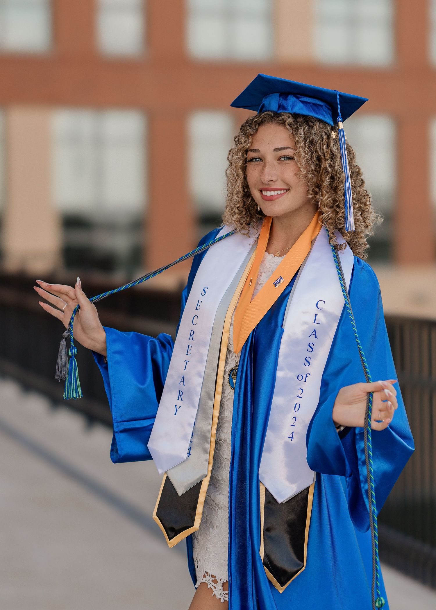 Graduate in blue cap and gown with curly hair poses for series of celebratory photos outdoors on graduation day.