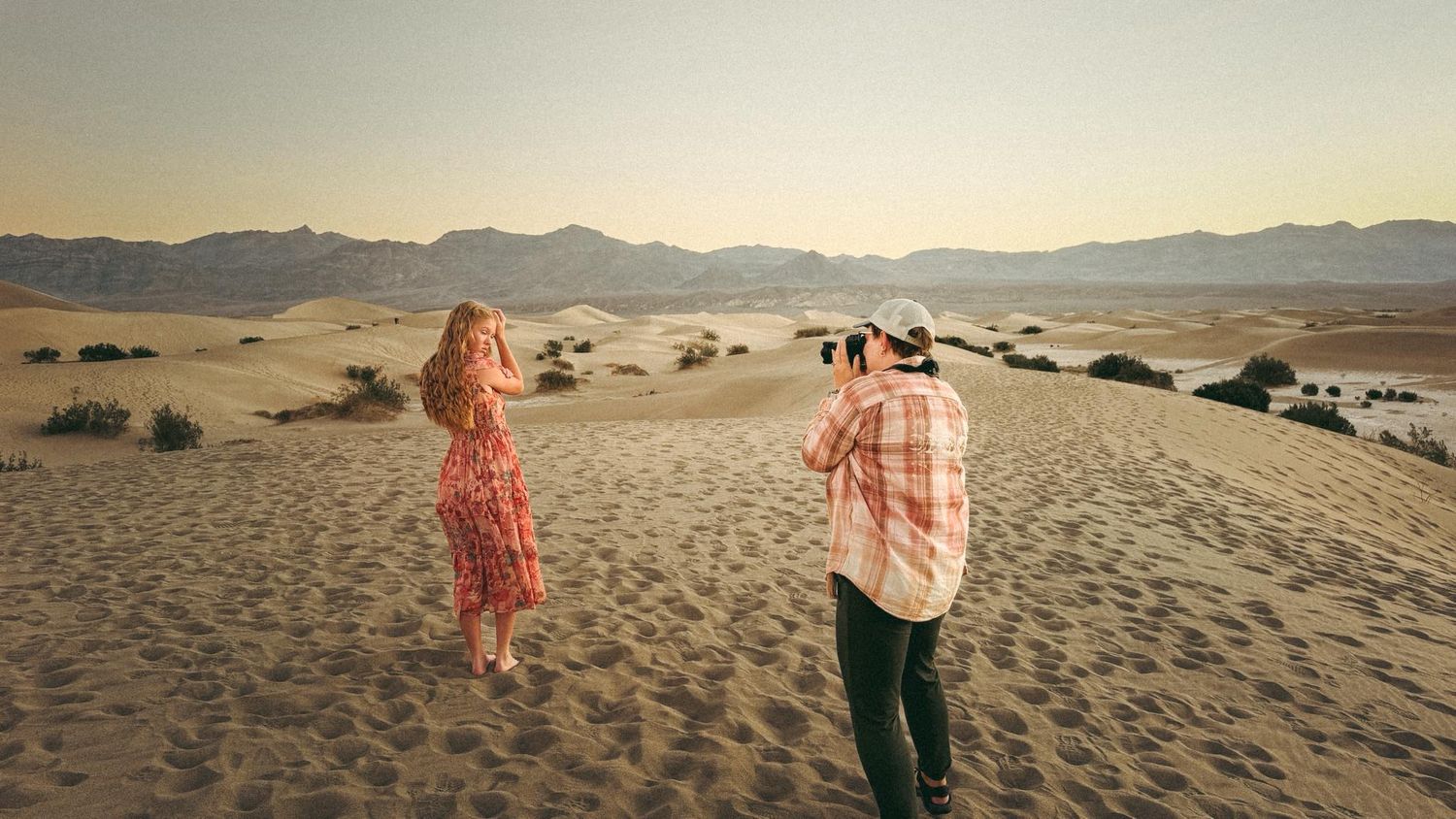 Couple photographing each other on sand dunes in desert landscape at sunset.
