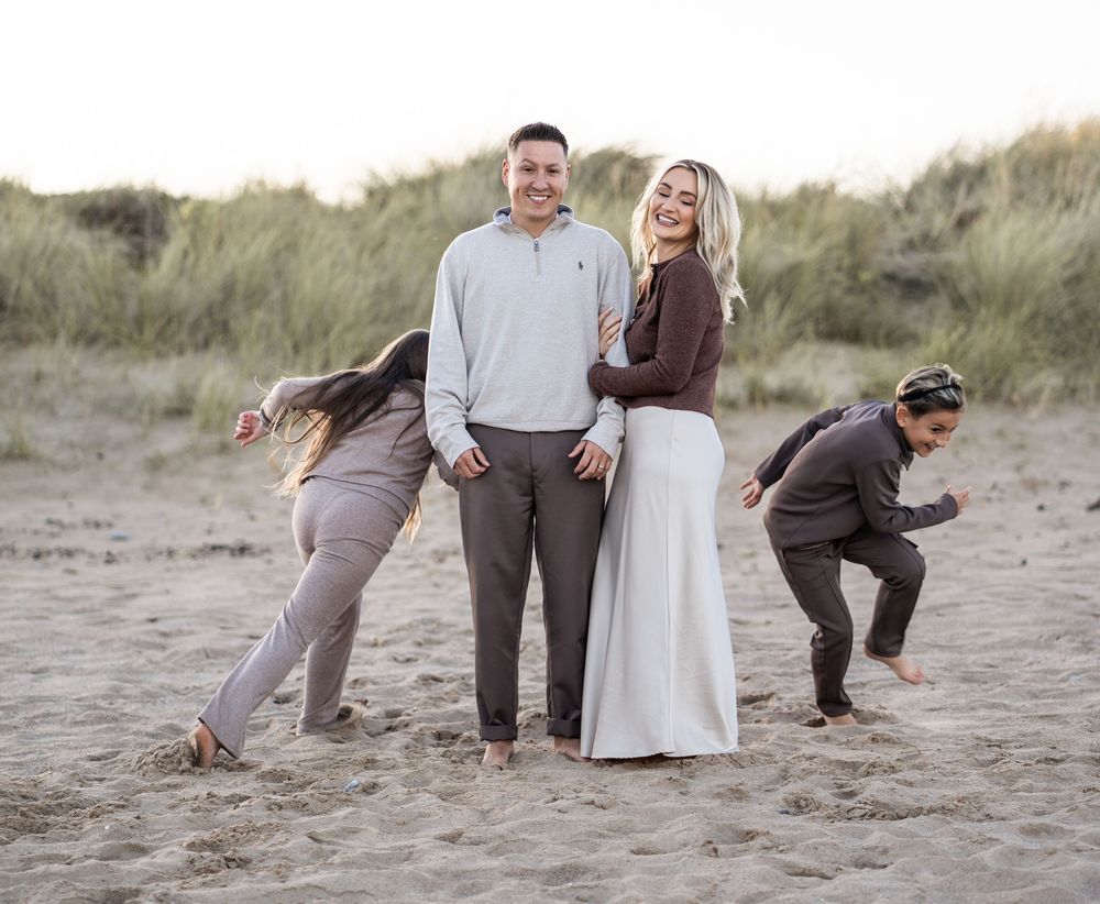 A playful group poses for wedding photos on a sandy beach with grassy dunes in the background.