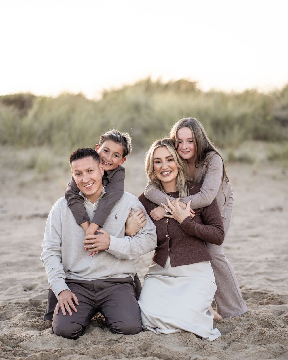 A family poses together sitting on a sandy beach during a casual outdoor photoshoot.