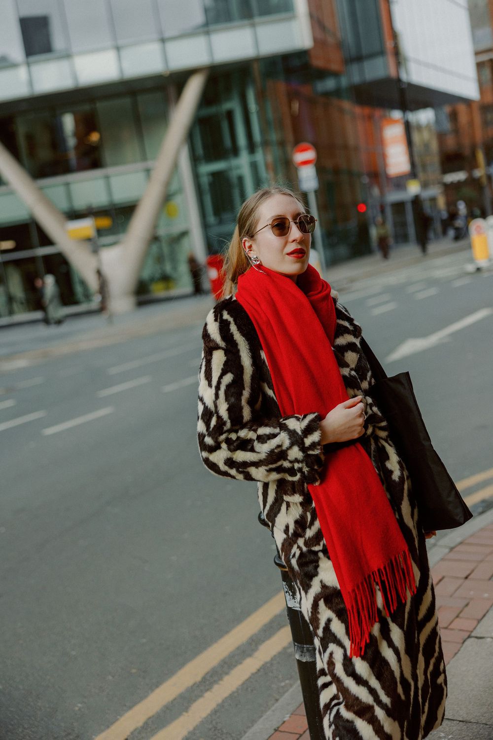 A person in a zebra print coat and bright red scarf poses on a city street corner with modern buildings in the background.