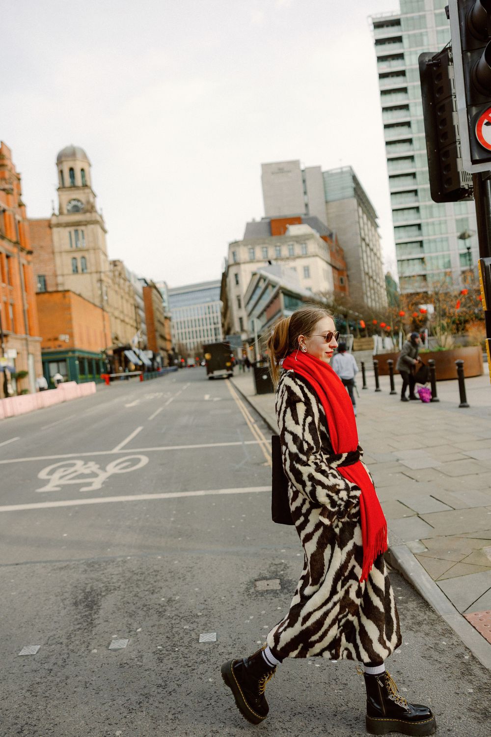 A person in a zebra print coat and red scarf poses on an urban street with tall buildings in the background.