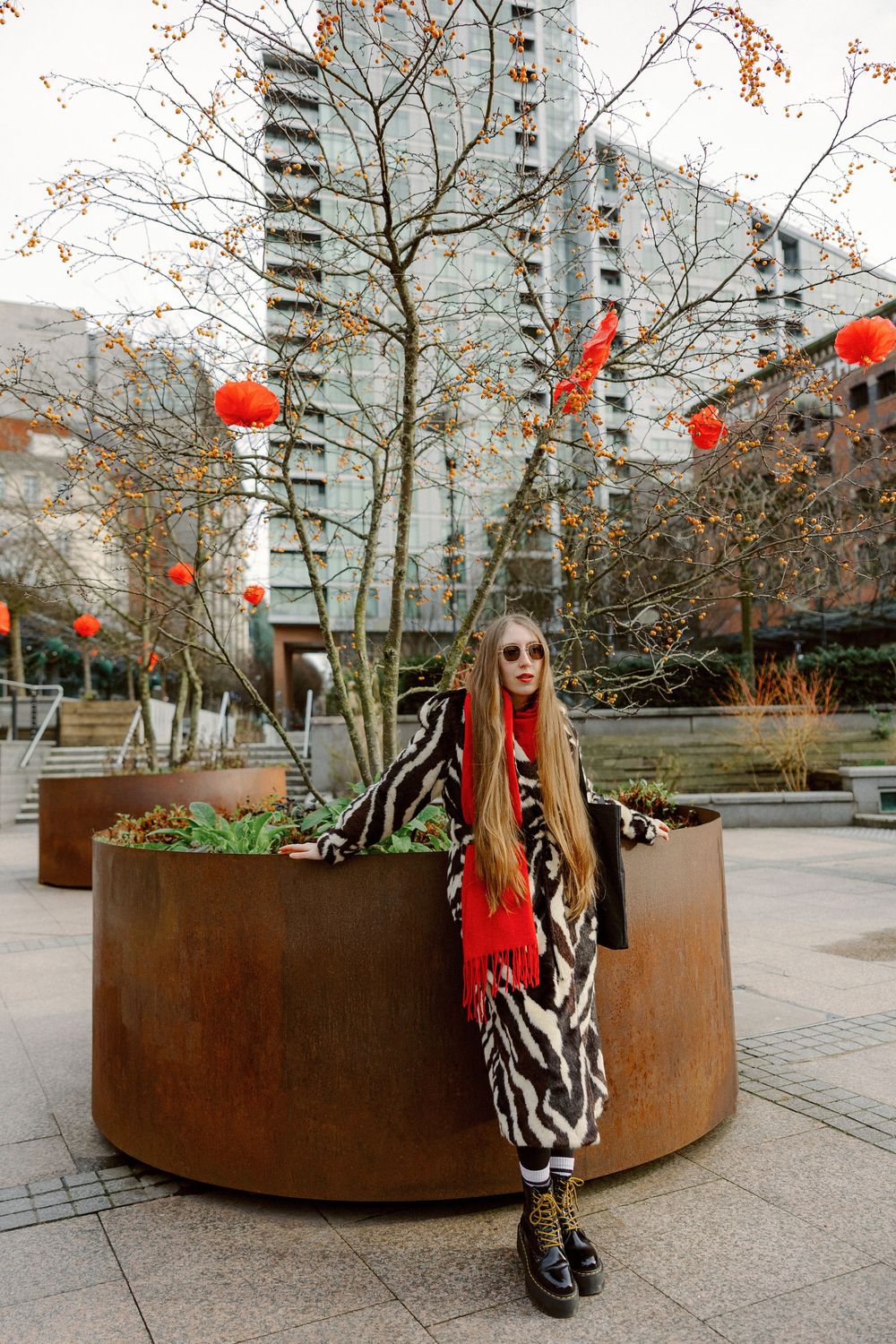 Person in zebra print coat and red sweater sits on curved metal planter with Chinese lanterns hanging above.