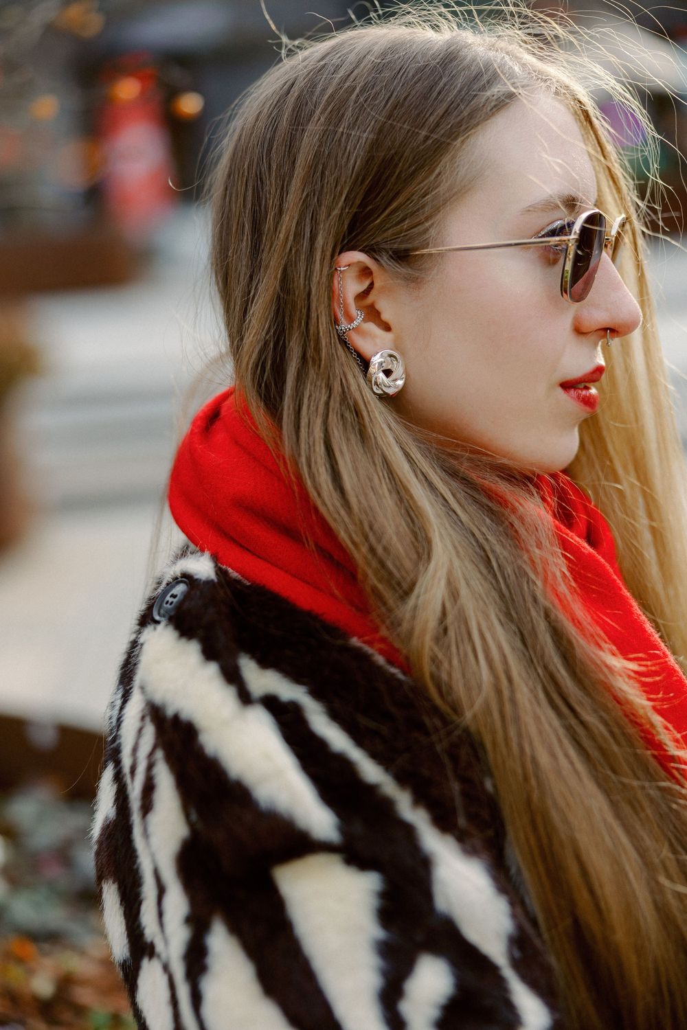 Stylish person wearing a black and white striped fur coat with a bright red scarf and sunglasses poses outdoors.